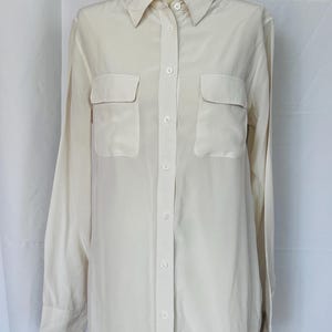 May include: A long-sleeved button-down shirt in a solid cream color. The shirt features a classic collar, a full-length front button placket, and two chest pockets with flaps. The fabric has a smooth, slightly lustrous finish and long sleeves.
