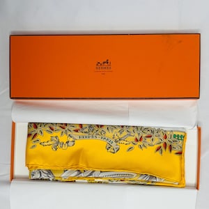 Peut inclure: Un foulard en soie jaune avec un motif floral et l'inscription "HERMES - PARIS" est présenté dans une boîte orange. L'écharpe a des accents rouges et verts. La boîte est ouverte, révélant l'écharpe à l'intérieur.