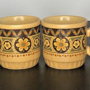 May include: Four beige ceramic mugs with a floral design. The mugs have a brown, green, and orange floral pattern around the middle. The handles and the inside of the mugs are beige.