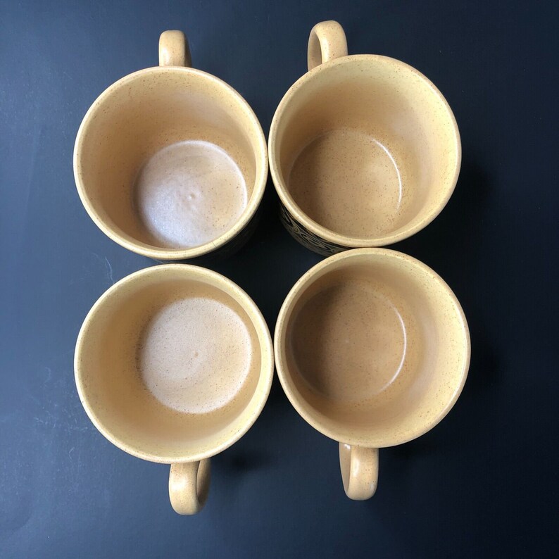 Op de afbeelding: Vier beige keramische mokken met handvatten, gerangschikt in een vierkant op een donker oppervlak. De mokken hebben een gespikkelde textuur en een lichtgekleurde binnenkant. De handvatten zijn gebogen en de mokken zijn leeg.