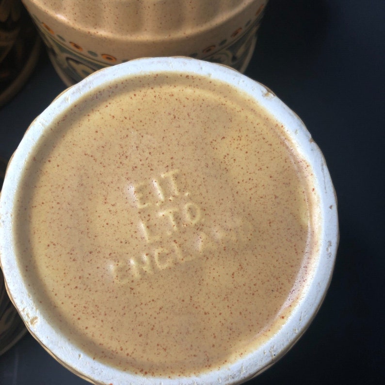 Op de afbeelding: Close-up van de bodem van een beige keramische container met een gespikkelde oppervlakte en een witte rand. De reli&euml;ftekst luidt "E.I.T. LTD. ENGLAND". De container maakt deel uit van een set, met een deel van een andere container zichtbaar op de achtergrond.