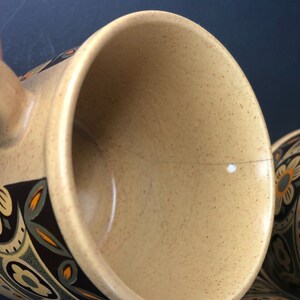 Op de afbeelding: Close-up van een beige keramische mok met een gespikkeld interieur. De mok heeft een decoratief bloemmotief in bruin, oranje en blauwgroen. De handgreep is zichtbaar aan de linkerkant. De mok maakt deel uit van een set.