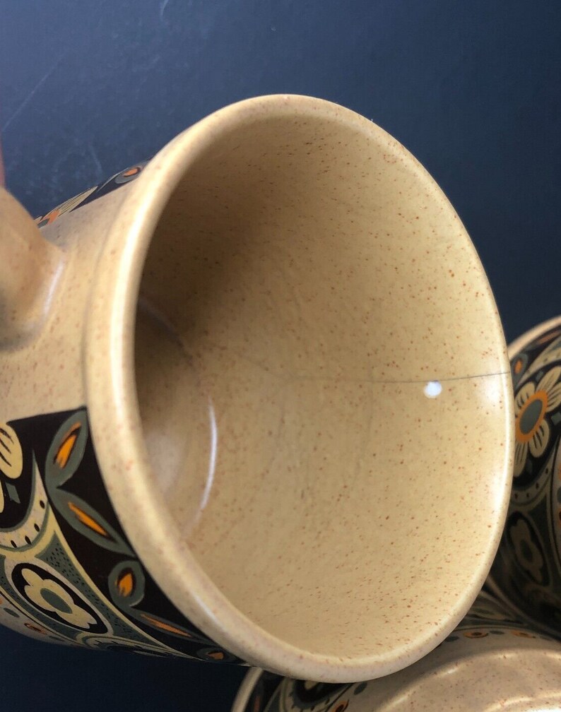 Op de afbeelding: Close-up van een beige keramische mok met een gespikkeld interieur. De mok heeft een decoratief bloemmotief in bruin, oranje en blauwgroen. De handgreep is zichtbaar aan de linkerkant. De mok maakt deel uit van een set.