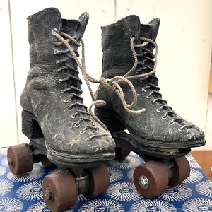 May include: A pair of vintage black roller skates with brown wheels and laces. The skates have a worn, distressed appearance, suggesting age and use. The laces are tied together. The skates are on a blue and white patterned surface.