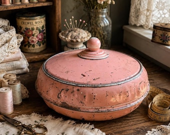 May include: A round, vintage-style metal tin with a distressed, dusty pink finish and a matching lid featuring a central knob. The container has visible signs of wear, including chipped paint and metallic edges, presenting a weathered appearance.