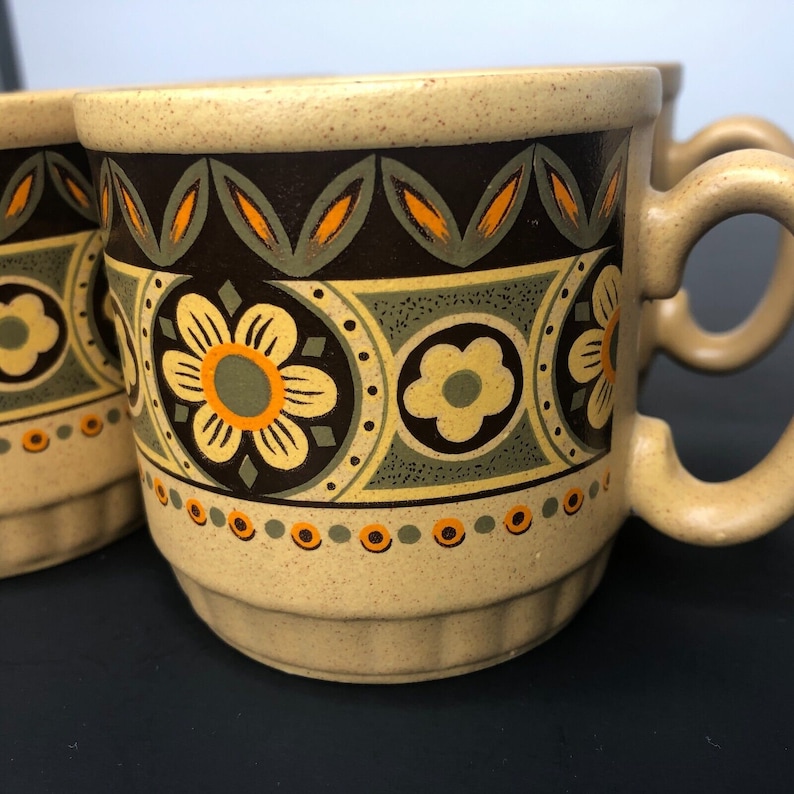 Op de afbeelding: Tan keramische mokken met een retro bloemenontwerp in bruin, groen en oranje. De mokken hebben een gespikkelde textuur en afgeronde handvatten. Het bloemenpatroon bevat grote bloemen en kleinere cirkelvormige accenten.