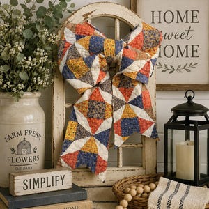 May include: A large decorative bow made from quilted fabric featuring a patchwork pattern of blue, orange, yellow, and white floral and geometric prints. The bow is draped over a white wooden arched window frame, creating a textured focal point.