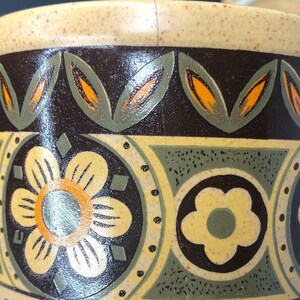 Op de afbeelding: Close-up van een keramische mok met een beige gespikkelde basis en een donkerbruine band met een bloemen- en bladerpatroon in geel, oranje en groen. De rand is beige.