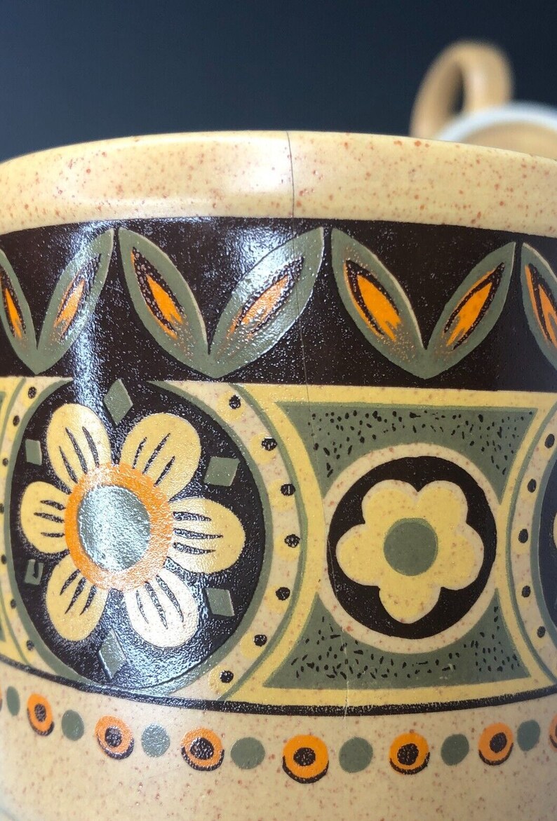 Op de afbeelding: Close-up van een keramische mok met een beige gespikkelde basis en een donkerbruine band met een bloemen- en bladerpatroon in geel, oranje en groen. De rand is beige.