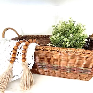 May include: A rectangular woven wicker basket with two arched handles, holding a white lace doily, wooden beads with tassels, and a small green plant. The basket has a natural brown color with accents of pink and green.