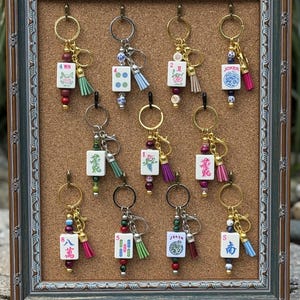 May include: A collection of eleven keychains featuring white mahjong tiles with various colorful symbols, numbers, and text including 'WIN', 'JOKER', and 'S'. Each keychain includes a metal ring, a small decorative tassel, and assorted beads.