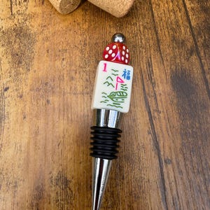 May include: A bottle stopper featuring a silver-toned metal base with black rubber rings, topped with a white mahjong tile displaying a red number one, a green boat, and a blue character, finished with a red die and a small silver bead.