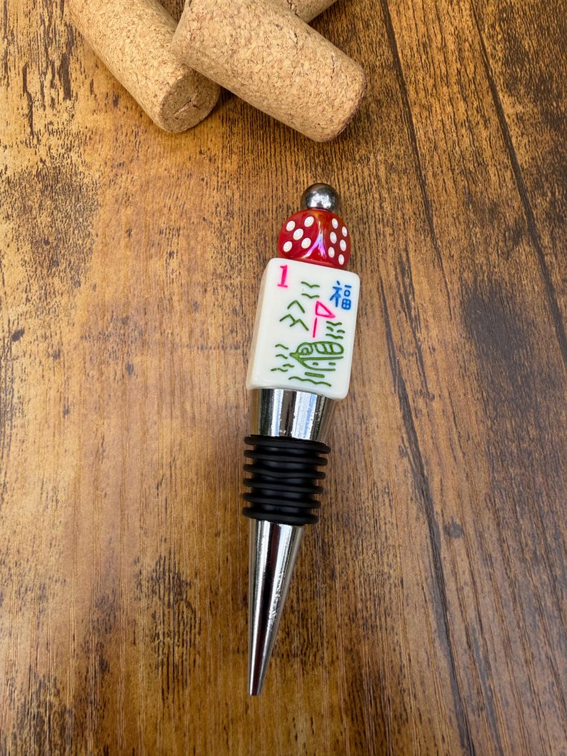 May include: A bottle stopper featuring a silver-toned metal base with black rubber rings, topped with a white mahjong tile displaying a red number one, a green boat, and a blue character, finished with a red die and a small silver bead.