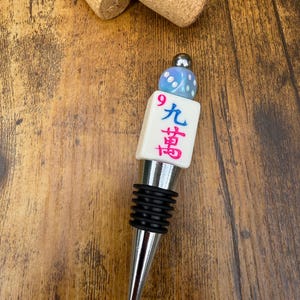 May include: A metal bottle stopper featuring a white mahjong tile with the Chinese character for nine and a red number 9, topped with a light blue iridescent die and a small silver bead. The stopper has a black ribbed rubber seal for a secure fit.