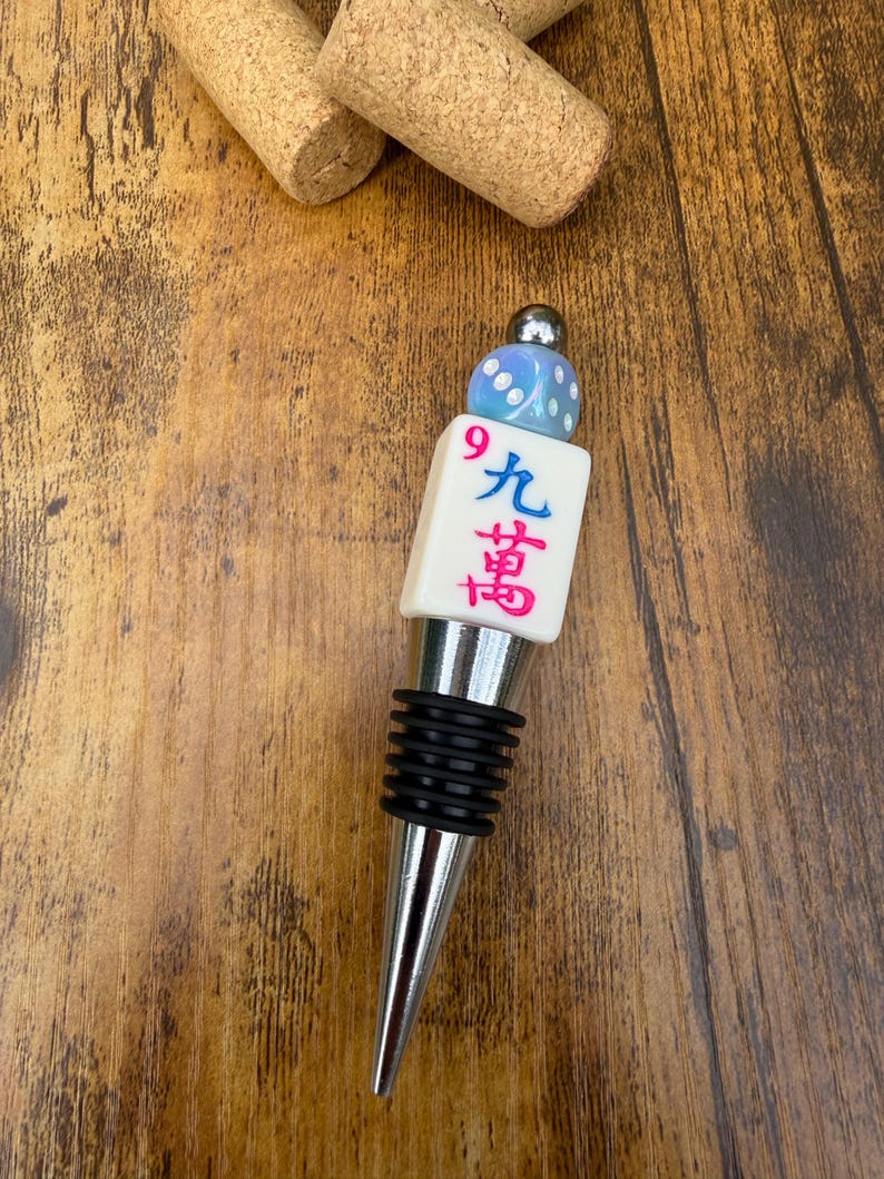 May include: A metal bottle stopper featuring a white mahjong tile with the Chinese character for nine and a red number 9, topped with a light blue iridescent die and a small silver bead. The stopper has a black ribbed rubber seal for a secure fit.