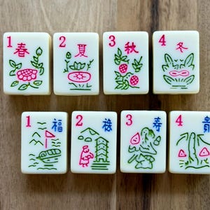 May include: Eight off-white rectangular mahjong tiles arranged in two rows. The top row features tiles numbered 1 through 4 with red numbers and green floral designs. The bottom row features tiles numbered 1 through 4 with red numbers and blue and green illustrations.