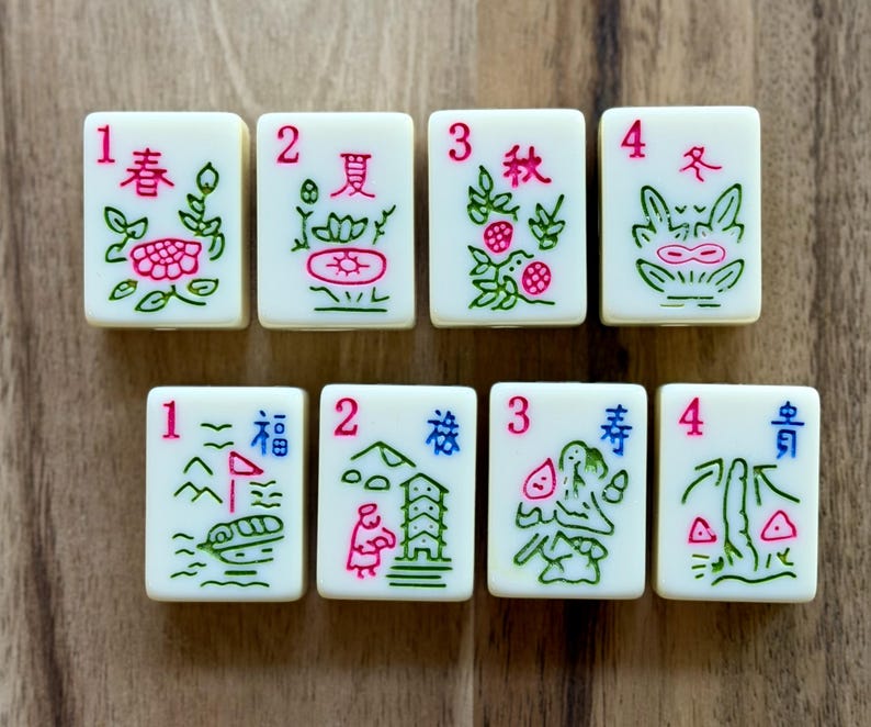 May include: Eight off-white rectangular mahjong tiles arranged in two rows. The top row features tiles numbered 1 through 4 with red numbers and green floral designs. The bottom row features tiles numbered 1 through 4 with red numbers and blue and green illustrations.