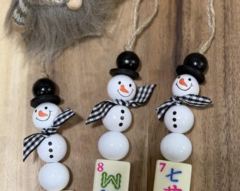 May include: Three snowman ornaments made of white wooden beads, black top hats, and black and white gingham scarves. Each snowman hangs from a jute twine loop and features a mahjong tile at the base, displaying a pink dragon, the letter M, or Chinese characters.