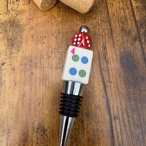 May include: A bottle stopper featuring a silver-toned metal base with black rubber rings, topped with a white square tile displaying a pink number 4 and four circular icons, finished with a red die and a small silver metal sphere.
