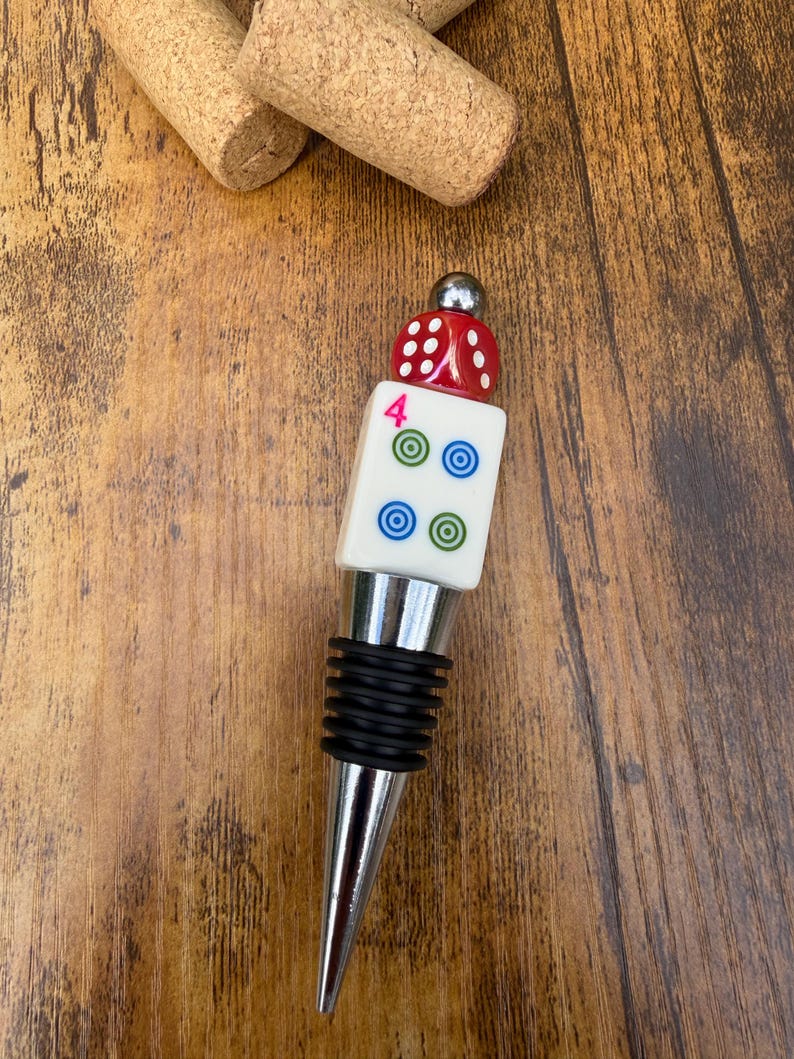 May include: A bottle stopper featuring a silver-toned metal base with black rubber rings, topped with a white square tile displaying a pink number 4 and four circular icons, finished with a red die and a small silver metal sphere.