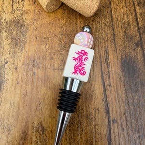 May include: A bottle stopper featuring a silver-toned metal base with black rubber rings, topped with a white rectangular mahjong tile displaying a pink dragon graphic, and finished with a small iridescent die and a silver bead.