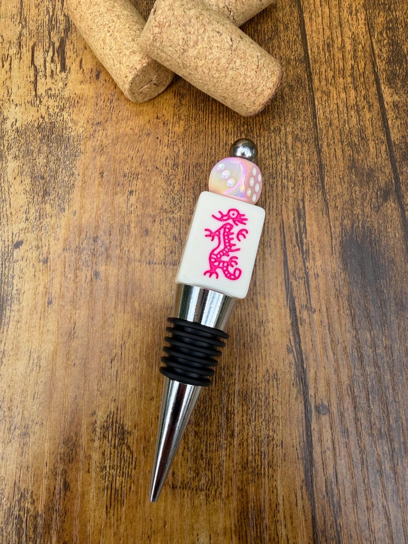 May include: A bottle stopper featuring a silver-toned metal base with black rubber rings, topped with a white rectangular mahjong tile displaying a pink dragon graphic, and finished with a small iridescent die and a silver bead.