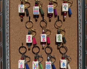 May include: A collection of eleven keychains featuring white mahjong tiles with various colorful symbols, numbers, and text including Joker, 3 AUT, and Chinese characters. Each keychain includes a gold metal ring, beads, and a small tassel.