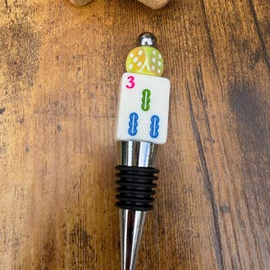 May include: A bottle stopper featuring a silver-toned metal base with black rubber rings, topped with a white rectangular Mahjong tile marked with a pink number 3 and green and blue symbols, finished with a yellow dice and a small silver bead.