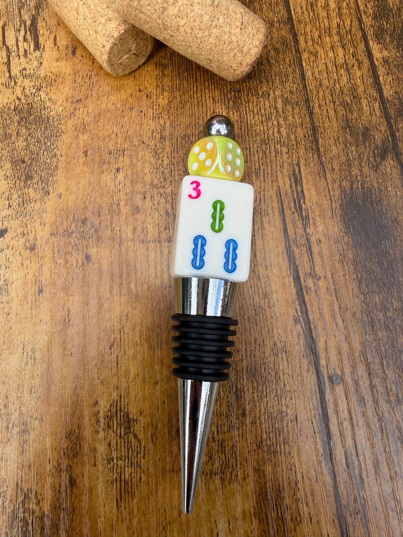 May include: A bottle stopper featuring a silver-toned metal base with black rubber rings, topped with a white rectangular Mahjong tile marked with a pink number 3 and green and blue symbols, finished with a yellow dice and a small silver bead.