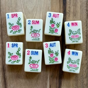 May include: Eight rectangular off-white Mahjong tiles featuring engraved floral designs in green and pink. Each tile displays a number and a seasonal abbreviation: 1 SPR, 2 SUM, 3 AUT, or 4 WIN, printed in blue and pink ink on the smooth surface.
