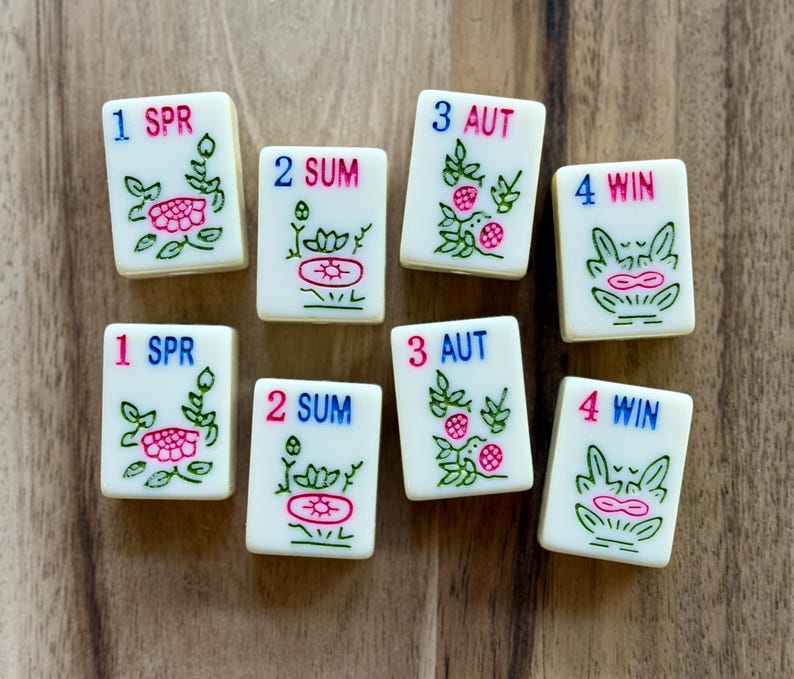 May include: Eight rectangular off-white Mahjong tiles featuring engraved floral designs in green and pink. Each tile displays a number and a seasonal abbreviation: 1 SPR, 2 SUM, 3 AUT, or 4 WIN, printed in blue and pink ink on the smooth surface.