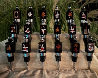 May include: A collection of twelve bottle stoppers featuring black Mahjong tiles as decorative toppers. Each tile displays unique red, white, and gold engraved symbols or characters. The stoppers have silver-toned metal bases with black rubber rings.