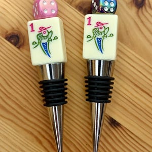 May include: Two bottle stoppers featuring a silver-toned metal base with black rubber rings. Each topper is topped with a white mahjong tile marked with the number 1 and a bird graphic, finished with a colorful die and a small silver sphere.