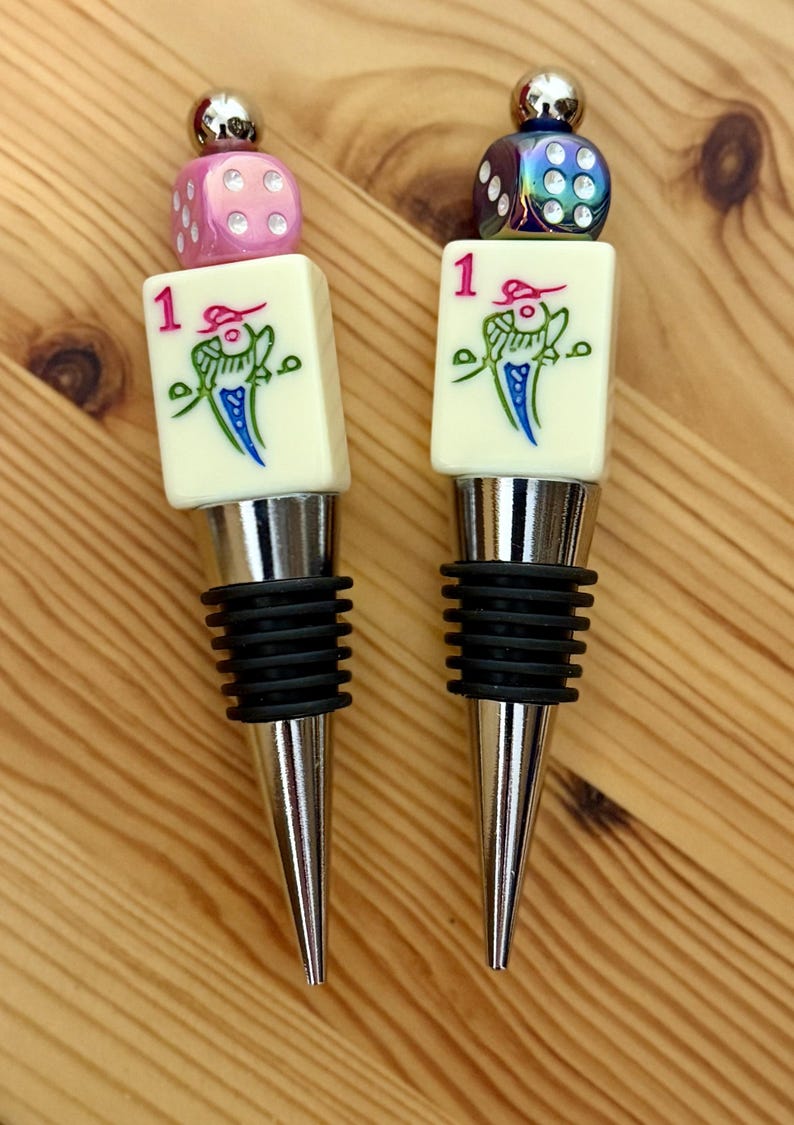 May include: Two bottle stoppers featuring a silver-toned metal base with black rubber rings. Each topper is topped with a white mahjong tile marked with the number 1 and a bird graphic, finished with a colorful die and a small silver sphere.