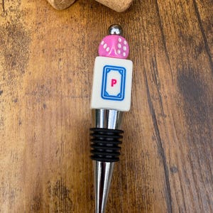 May include: A bottle stopper featuring a silver-toned metal base with black rubber rings, topped with a white rectangular tile displaying a blue border and the letter P in pink, finished with a pink die and a small silver metal sphere.