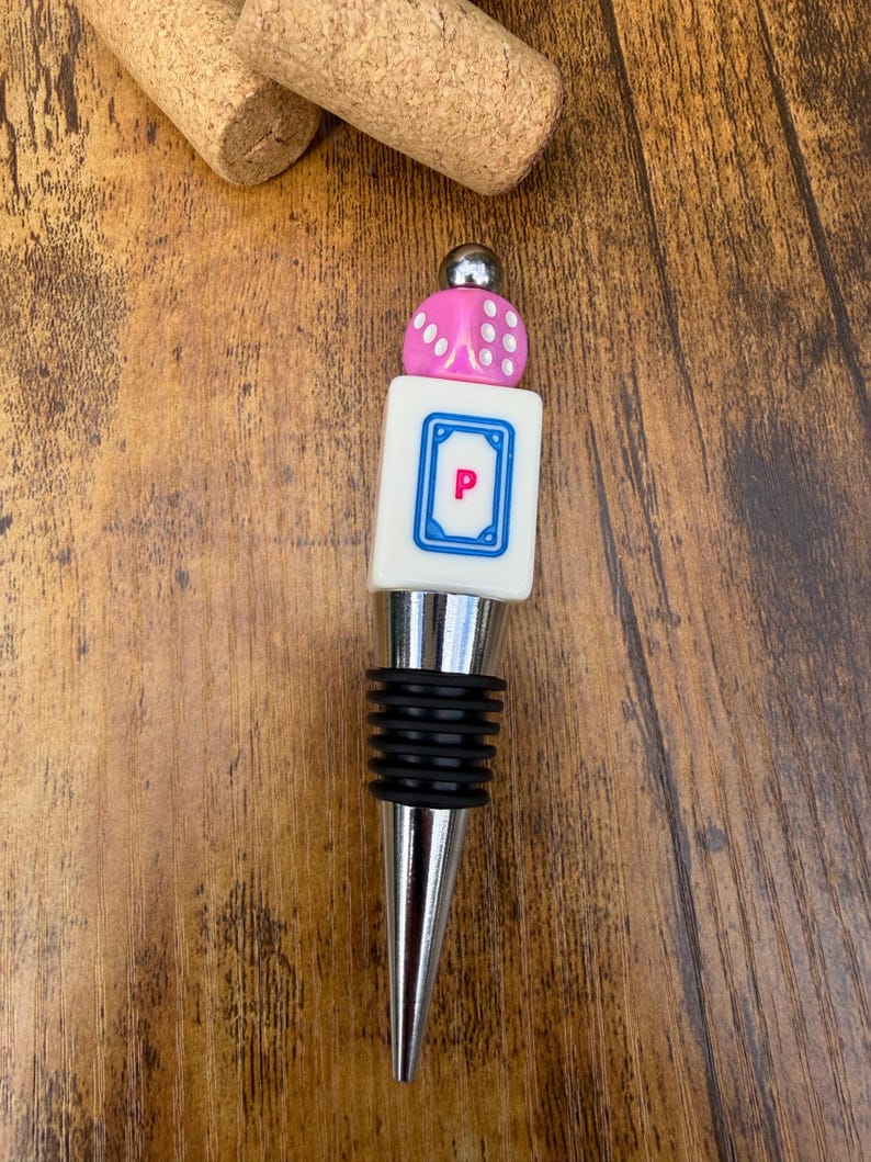 May include: A bottle stopper featuring a silver-toned metal base with black rubber rings, topped with a white rectangular tile displaying a blue border and the letter P in pink, finished with a pink die and a small silver metal sphere.