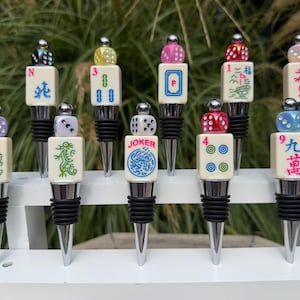 May include: Ten bottle stoppers with silver-toned metal bases and black rubber rings. Each stopper features a cream-colored mahjong tile topped with a small, colorful die. The tiles display various symbols, numbers, and the word JOKER in red ink.