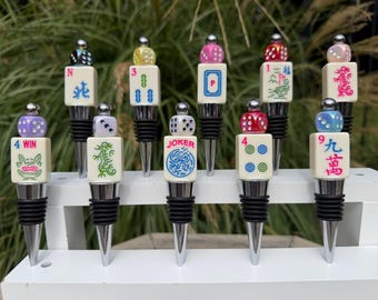 May include: A collection of ten bottle stoppers featuring cream-colored mahjong tiles topped with colorful dice. Each stopper has a silver-toned metal base with black rubber rings. The tiles display various symbols, numbers, and the word JOKER.