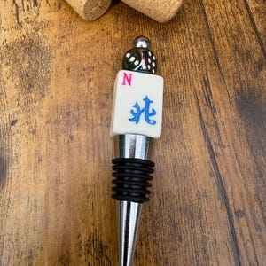 May include: A bottle stopper featuring a metallic silver base with black rubber rings, topped with a white Mahjong tile displaying a pink letter N and a blue character, finished with a small green translucent die at the very top.