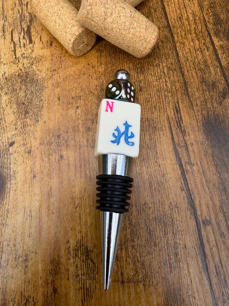 May include: A bottle stopper featuring a metallic silver base with black rubber rings, topped with a white Mahjong tile displaying a pink letter N and a blue character, finished with a small green translucent die at the very top.