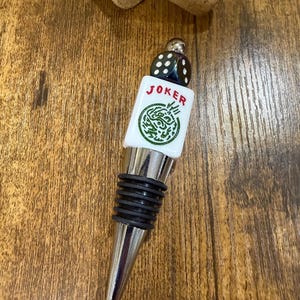 May include: A bottle stopper featuring a silver-toned metal base with black rubber rings, topped with a white rectangular tile displaying the red text JOKER above a green circular graphic, finished with a small black die at the very top.