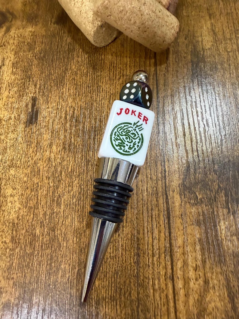 May include: A bottle stopper featuring a silver-toned metal base with black rubber rings, topped with a white rectangular tile displaying the red text JOKER above a green circular graphic, finished with a small black die at the very top.