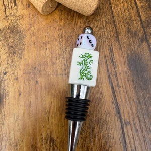 May include: A bottle stopper featuring a silver-toned metal base with black rubber rings, topped with a white rectangular Mahjong tile displaying a green dragon design, a small iridescent die, and a polished silver-toned metal bead.