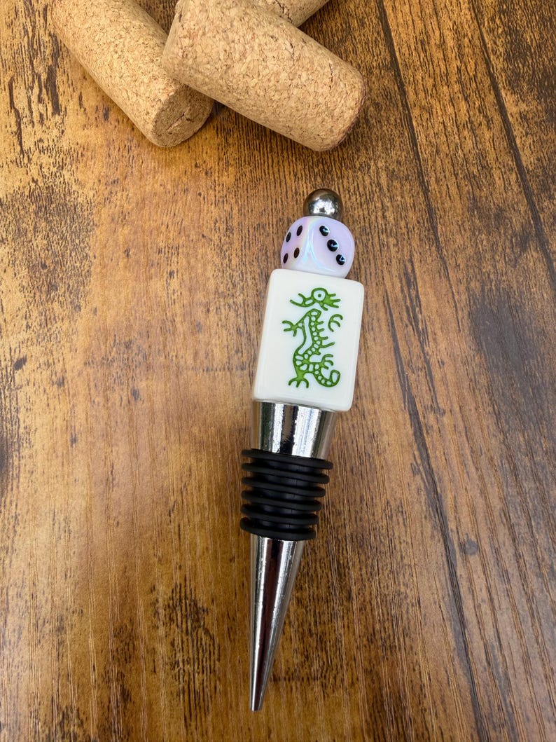 May include: A bottle stopper featuring a silver-toned metal base with black rubber rings, topped with a white rectangular Mahjong tile displaying a green dragon design, a small iridescent die, and a polished silver-toned metal bead.