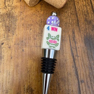 May include: A bottle stopper featuring a silver-toned metal base with black rubber rings, topped with a white rectangular block printed with the text 4 WIN and a green graphic, finished with a purple die bead and a small silver metal sphere.