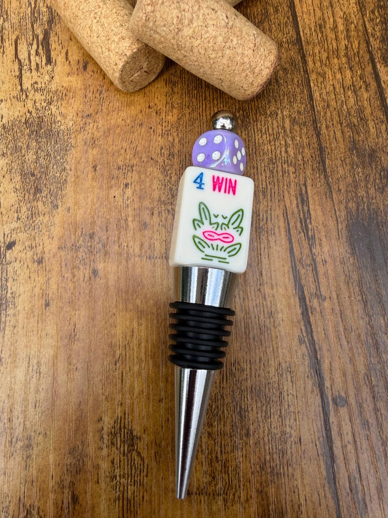May include: A bottle stopper featuring a silver-toned metal base with black rubber rings, topped with a white rectangular block printed with the text 4 WIN and a green graphic, finished with a purple die bead and a small silver metal sphere.