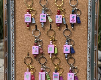 May include: Eleven keychains featuring pink Mahjong tiles with various symbols and characters. Each keychain includes a metal ring, a lobster clasp, a small colorful tassel, and decorative beads in assorted colors and patterns, all displayed on a cork board.
