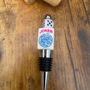 May include: A bottle stopper featuring a silver-toned metal base with black rubber rings, topped with a white rectangular block labeled JOKER in pink text above a blue circular graphic, and a small white six-sided die with black pips.