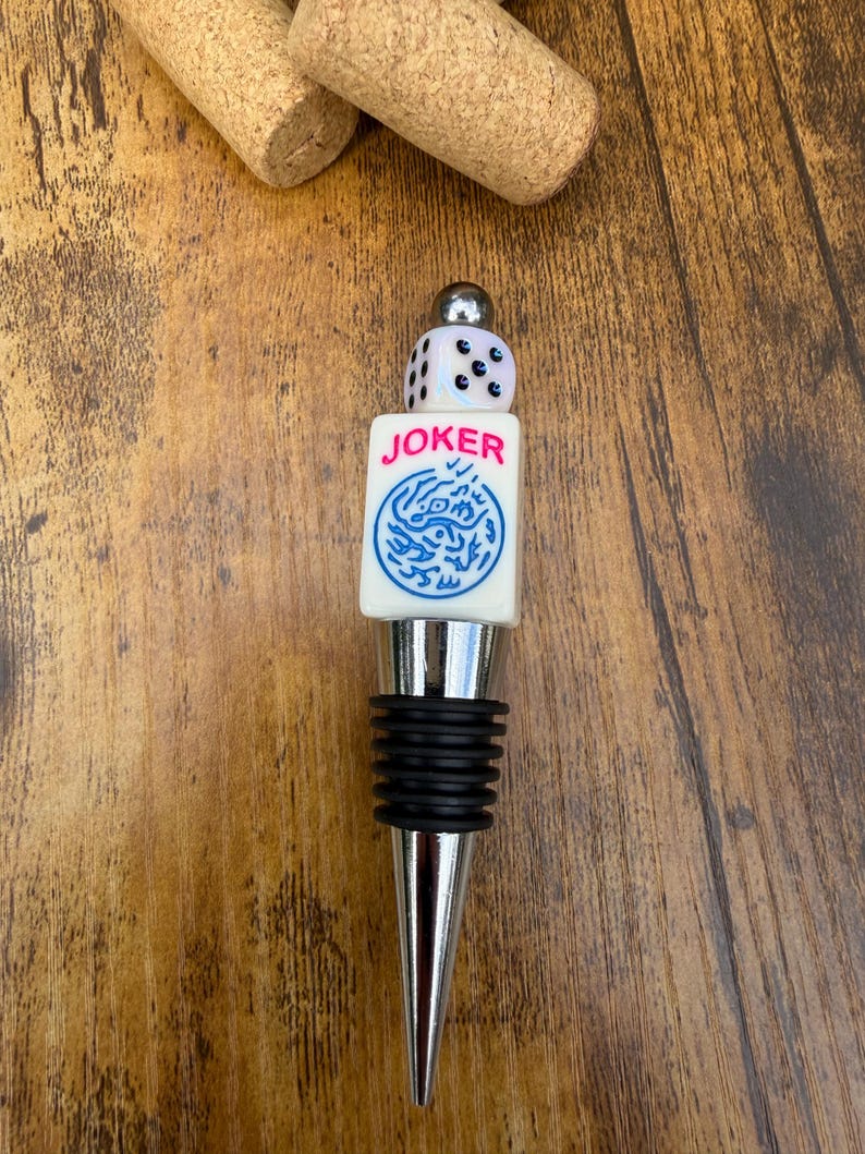 May include: A bottle stopper featuring a silver-toned metal base with black rubber rings, topped with a white rectangular block labeled JOKER in pink text above a blue circular graphic, and a small white six-sided die with black pips.