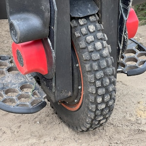 May include: A black electric unicycle with a large knobby tire, red footrests, and black metal foot pedals. The unicycle is on a sandy surface.